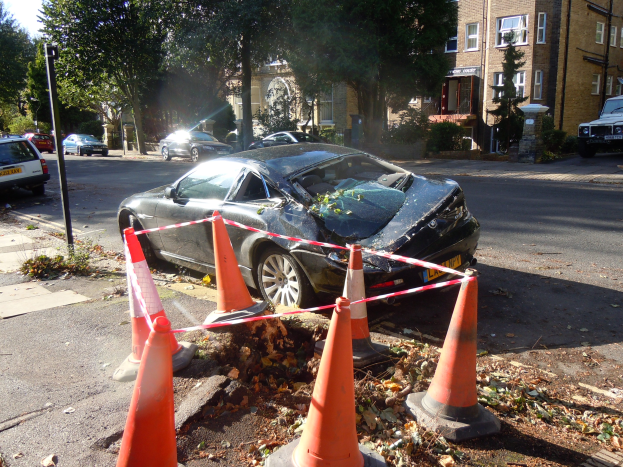 Ein besch├Ądigt auf der Stra├če stehendes Auto mit orangefarbenen Absperrbaken drumherum, B├Ąume und Geb├Ąude im Hintergrund unter einem klaren blauen Himmel.