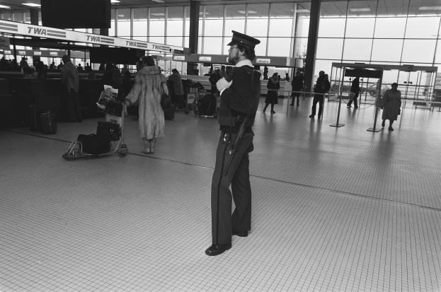 Polizist in Uniform mit Waffe in einem Flughafen-Terminal umgeben von Menschen und Gepäck, mit Gepäckwagen, Deckenleuchten, Glaswänden und Informationsanzeigen im Hintergrund.