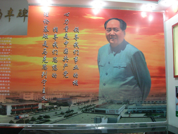 Großes Mao-Zedong-Plakat an einer Gebäudewand, beleuchtet von Lampen, mit einem Foto Rahmen und Text rechts daneben.