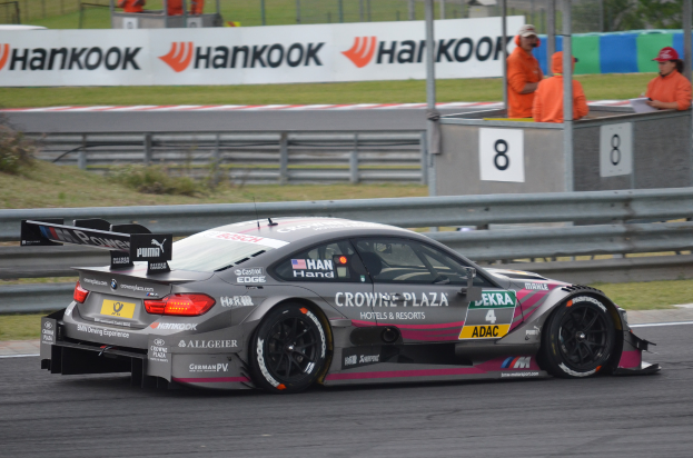Ein BMW M4 DTM-Rennwagen auf einer Rennstrecke, umgeben von einem Metallzaun, Gras, Pflanzen, einer Gruppe von Menschen, einem Schild mit Text, Pfosten, einem Metallrahmen, Bäumen und einem bewölkten Himmel.