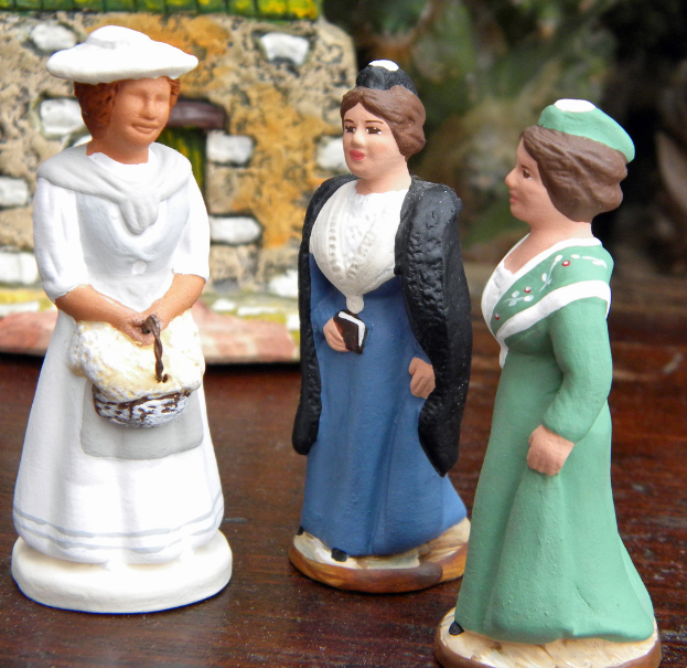 Drei Skulpturenpuppen sind auf einer Holzplatte mit Spielzeughäusern im Hintergrund angeordnet.