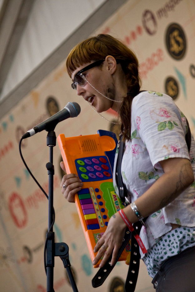 Eine Frau in einem wei├čen Hemd und Sonnenbrille singt in ein Mikrofon auf einem St├Ąnder, h├Ąlt eine Tastatur und hat ein Plakat an der Wand im Hintergrund.