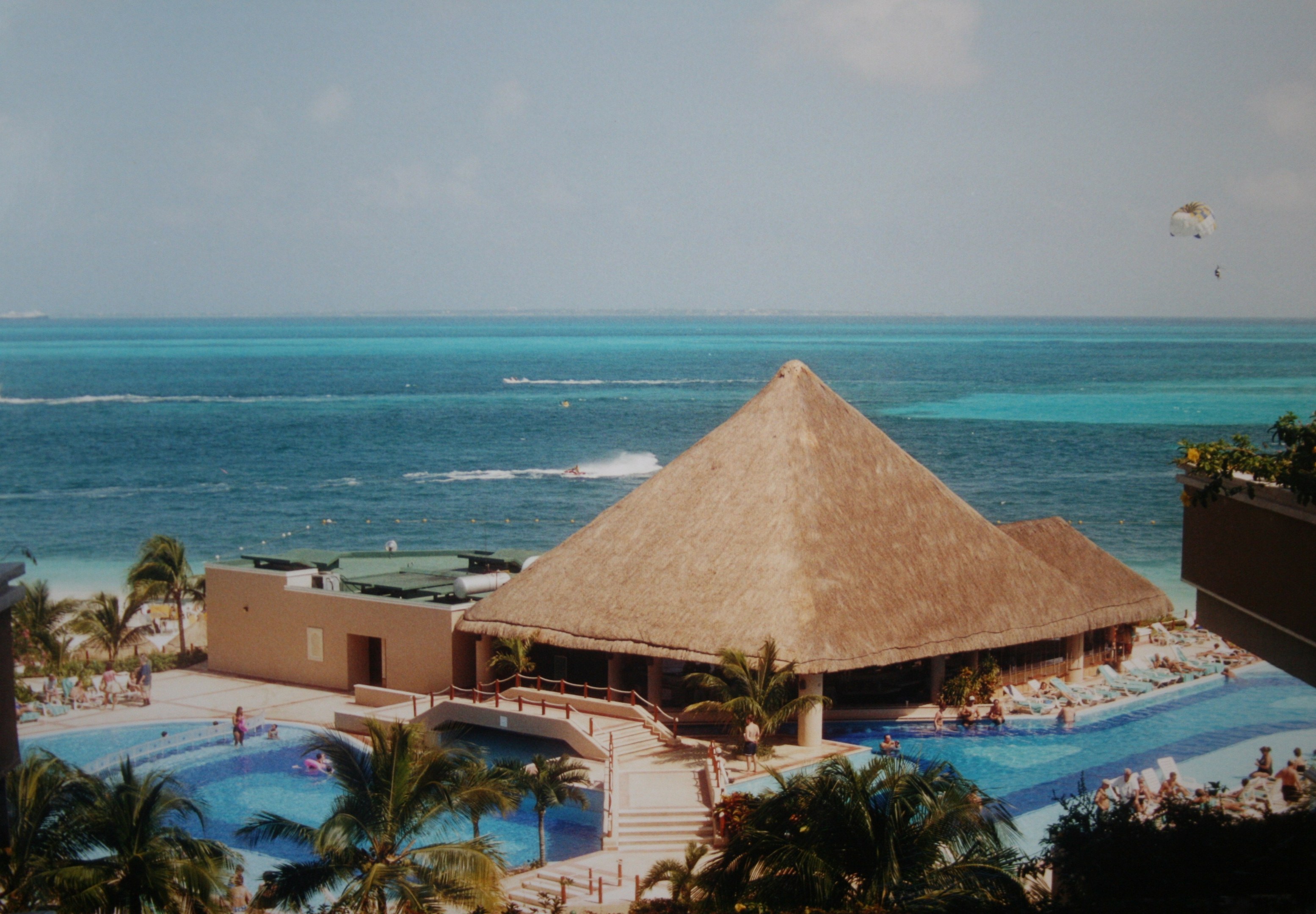 Resort mit einem gro├čen Schwimmbad umgeben von Palmen, Hütten und Menschen, mit einem Ozean und einem bew├Âlktem Himmel im Hintergrund, das einen Fallschirm in der Luft zeigt.