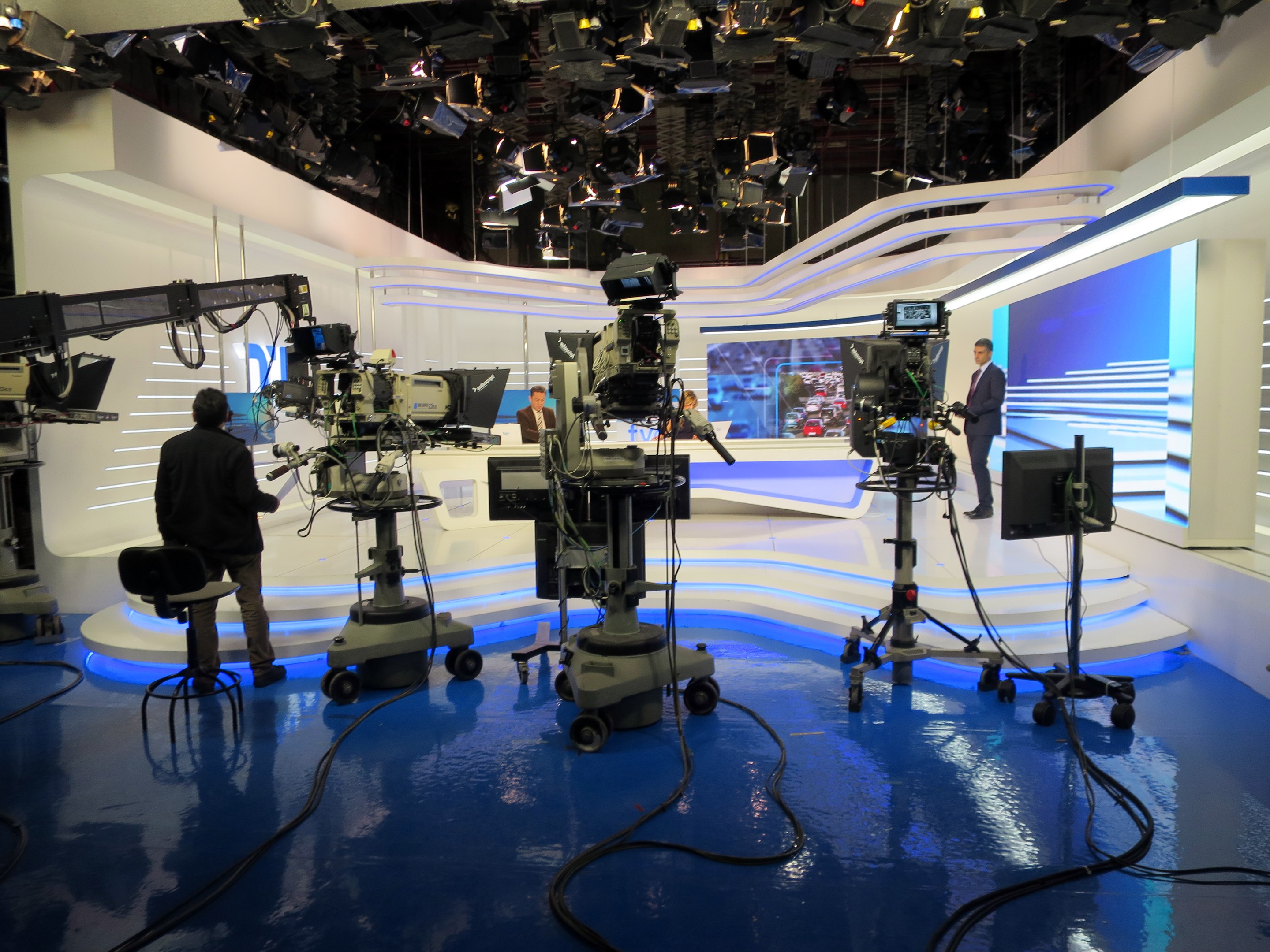 Ein TV-Studio mit mehreren Kameras auf dem Boden, einer sitzenden Person im Vordergrund und einigen stehenden Personen im Hintergrund, sowie an der Decke montierten Scheinwerfern für eine Livesendung.