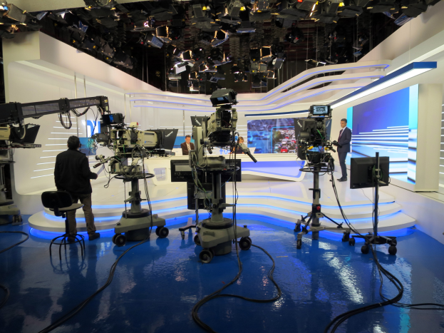 Ein TV-Studio mit mehreren Kameras auf dem Boden, einer sitzenden Person im Vordergrund und einigen stehenden Personen im Hintergrund, sowie an der Decke montierten Scheinwerfern für eine Livesendung.