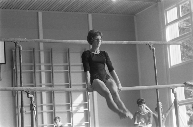 Ein Schwarz-Weiß-Foto einer Frau, die an einem Gymnastikgerät in einem Turnsaal trainiert, mit einigen Menschen, einer Wand, einer Leiter und einem Fenster im Hintergrund.