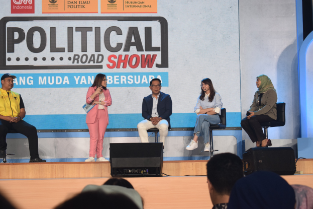 Eine Gruppe von Menschen sitzt auf einer Bühne mit einer Frau, die ein Mikrofon in der Mitte hält, einem Publikum im Vordergrund und einem Banner mit der Aufschrift "Politischer Straßenumzug 2019" im Hintergrund.