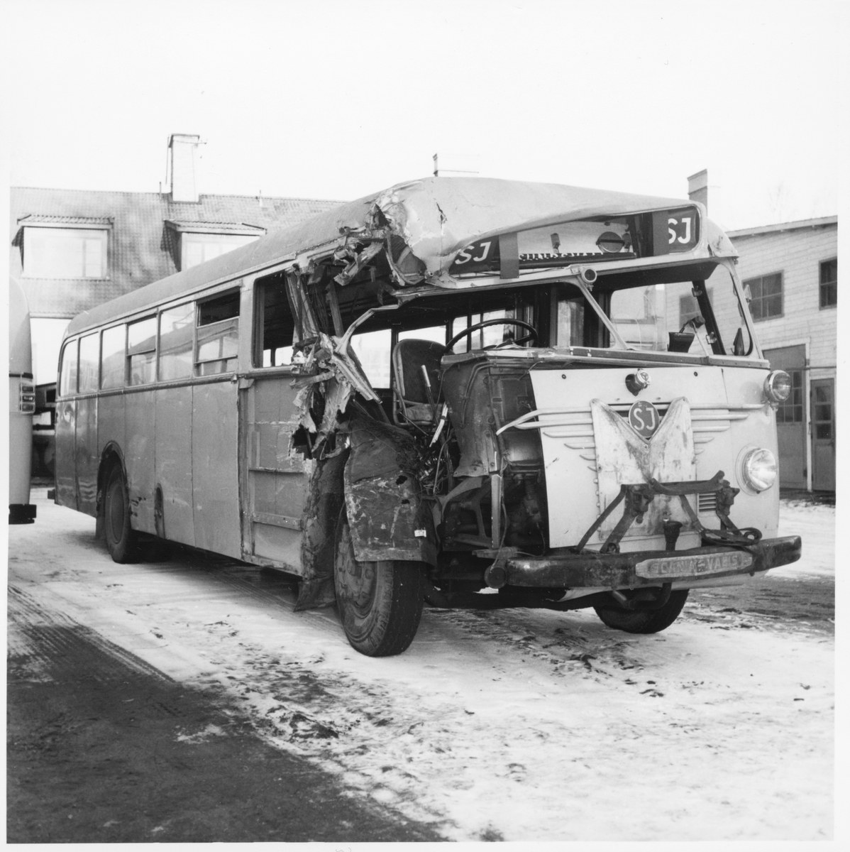 Schwarzes und weißes Foto eines schwerbeschädigten Busses mit eingedrückter Front und zersplitterten Fenstern an der Seite einer Straße, mit Gebäuden im Hintergrund.
