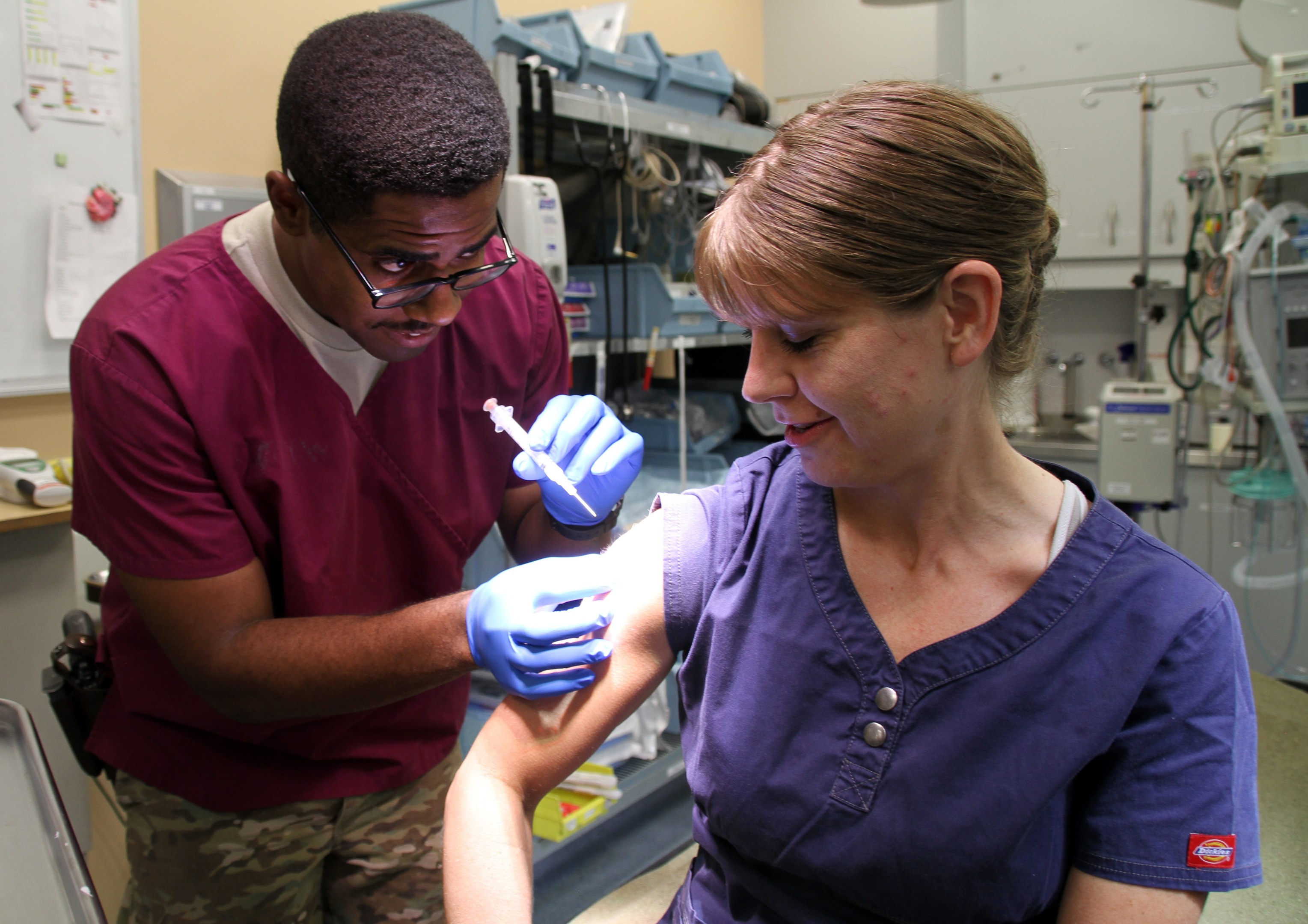 Ein Mann und eine Frau in Kitteln erhalten Impfungen von einer Krankenschwester, wobei der Mann eine Spritze hält und die Frau Handschuhe trägt, in einer medizinischen Einrichtung mit Geräten und Papieren an der Wand.
