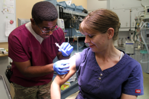 Ein Mann und eine Frau in Kitteln erhalten Impfungen von einer Krankenschwester, wobei der Mann eine Spritze hält und die Frau Handschuhe trägt, in einer medizinischen Einrichtung mit Geräten und Papieren an der Wand.