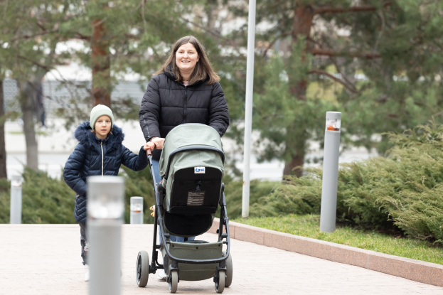 Eine Frau schiebt einen Kinderwagen mit einem Kind eine von Pfosten, Gras, Pflanzen und Bäumen gesäumte Gehweg.