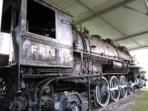 Ein Zug ist auf Schienen in der Mitte zu sehen, darunter Gras und darüber eine Decke sowie Stangen.