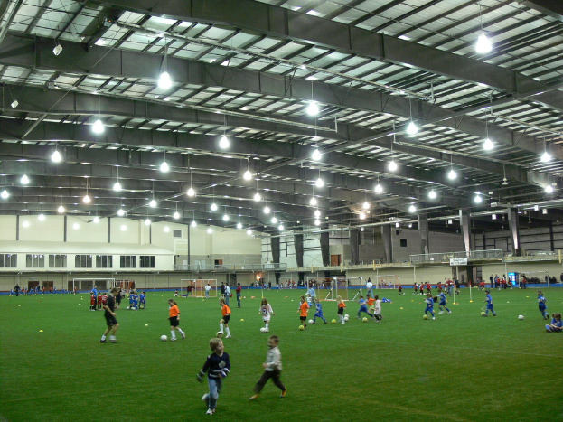 Eine Halle mit einer Gruppe von Menschen, die mit einem Fußball auf dem Boden spielt, Torpfosten und Beleuchtung auf dem Dach.