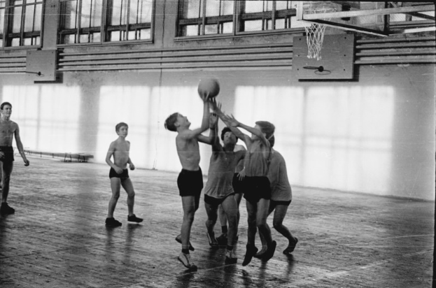 Eine Gruppe junger Männer, die Basketball in einer Turnhalle spielen, mit einer Wand im Hintergrund und Fenstern oben, dargestellt in Schwarz-Weiß.