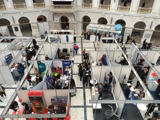 Große Gruppe von Besuchern auf einer internationalen Fachmesse mit Ständen, Bannern, Tischen, Stühlen und Ballons in einem Raum mit architektonischen Bögen, Säulen und einem Bildschirm im Hintergrund.