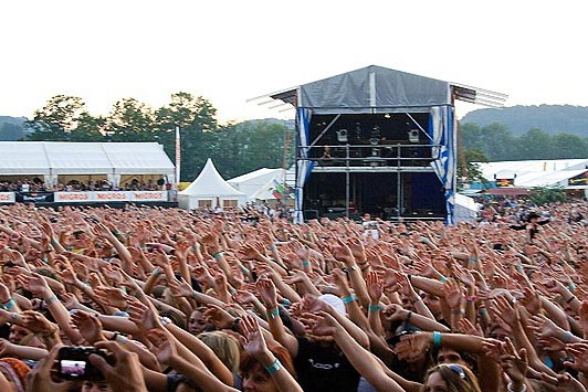 Eine große Menschenmenge auf einem Musikfestival mit erhobenen Händen und Mobiltelefonen, eine Bühne mit Performern im Hintergrund, weiße Zelte, Bäume und einen klaren blauen Himmel.