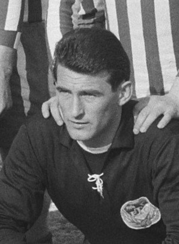 Schwarzes und weißes Foto eines Mannes, der auf einem grünen Fußballfeld sitzt und ein T-Shirt und eine Kette trägt, mit zwei Personen hinter ihm.