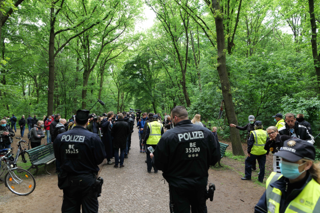 Eine Gruppe von Polizisten steht vor einer Menschenmenge, einige tragen Mützen und Masken, mit Fahrrädern und einer Bank im Vordergrund und Bäumen und Himmel im Hintergrund, während sie an einer Anti-Terror-Demonstration in Berlin teilnehmen.
