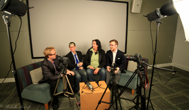 Eine Gruppe von drei Personen sitzt auf einem Sofa vor einem Projektionsschirm, wobei eine Person ein Mikrofon hält, während eine Person in einem schwarzen Mantel vor ihnen sitzt, und Kameras und Beleuchtungsausrüstung auf beiden Seiten positioniert sind.
