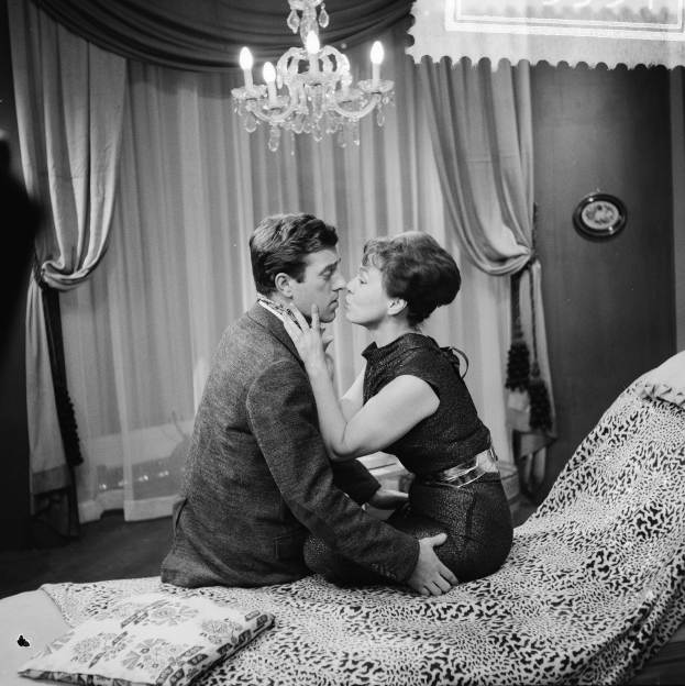 Ein Mann und eine Frau auf einem Bett sitzend, die sich umarmen und küssen, mit Kissen, einer Decke und einem Kronleuchter im Raum und Vorhängen im Hintergrund.