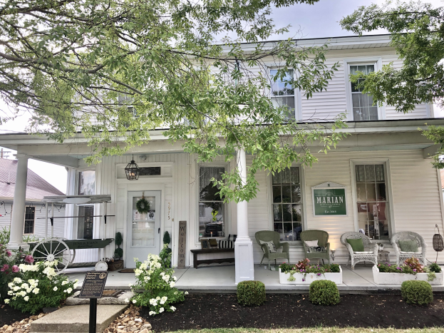 Das Marian Inn, ein weißes Haus mit einer Veranda, Säulen, Fenstern und einer Tür, steht vor einem Baum mit einem Tisch und Stühlen auf der Veranda, umgeben von Pflanzen, Blumen, Gras, Steinen und einer Gruppe von Bäumen unter einem bewölkten Himmel.
