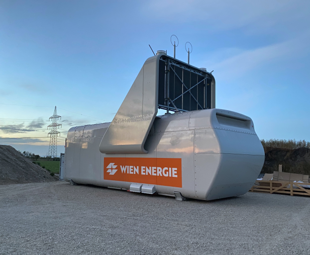 Eine große weiße und orangefarbene Windkraftanlage steht auf einem Schotterfeld umgeben von Bäumen und einem Turm im Hintergrund, mit ein paar Gegenständen rechts auf dem Boden und dem Himmel oben sichtbar.