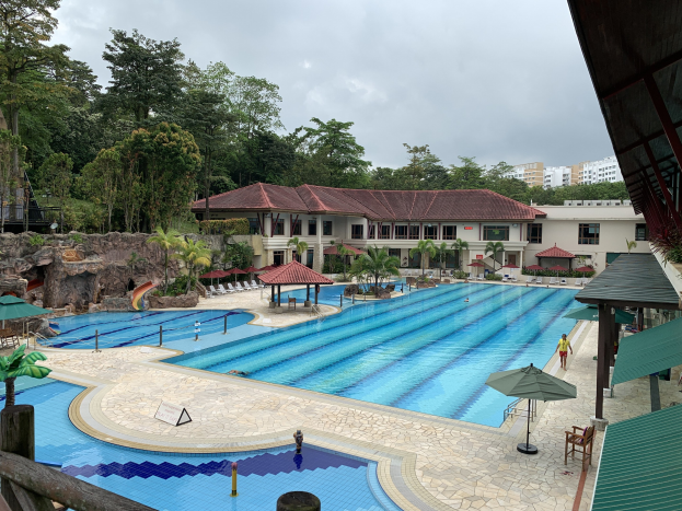 Großes Schwimmbecken im Ritz-Carlton, Kota Kinabalu, mit Menschen, Sonnenschirmen, Stühlen, Tischen, Pflanzen, Bäumen, Felsen, Gebäuden und einem Schuppen auf der rechten Seite sowie einem sichtbaren Himmel im Hintergrund.