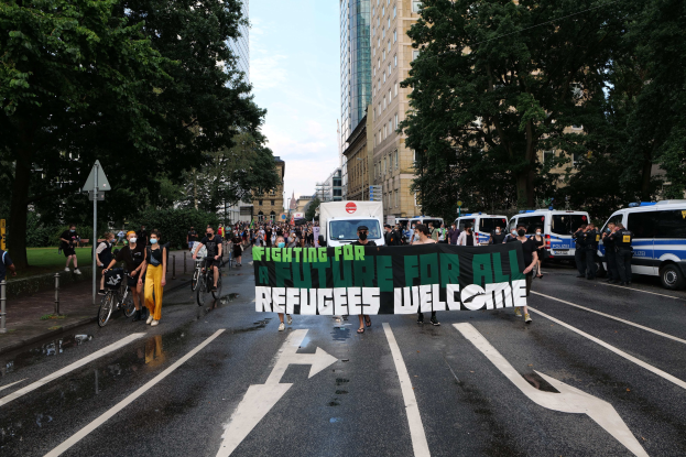 Eine Gruppe von Menschen, die eine Straße entlanggehen und ein 'Refugees Welcome'-Schild halten, mit parkenden Fahrzeugen, Radfahrern und Bäumen an der Straße und einem sichtbaren Himmel.