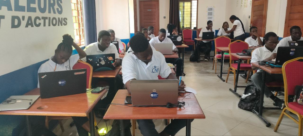 Eine Gruppe von Menschen sitzt an Tischen mit Laptops in einem Klassenzimmer, mit Taschen auf dem Boden und Text an der linken Wand, was auf einen Online-Kurs in Ghana hinweist.