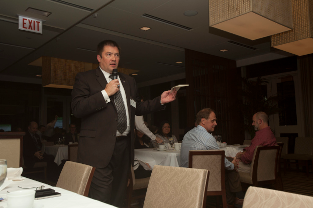 Ein Mann steht an einem Mikrofon und spricht zu einem sitzenden Publikum, mit Tassen auf den Tischen und einem Schild an der Decke.