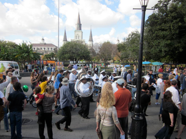 Eine Gruppe von Menschen auf einer Straße, einige spielen Musikinstrumente, umgeben von Gebäuden, Bäumen, Pflanzen, Zäunen, Fahrzeugen und Masten mit Laternen.