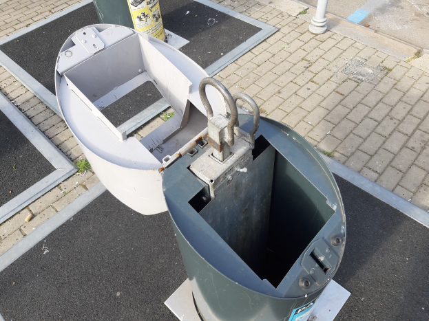 Ein Metallmüllcontainer an der Straßenseite mit einem Metallobjekt im Vordergrund und einem Pfahl im Hintergrund.