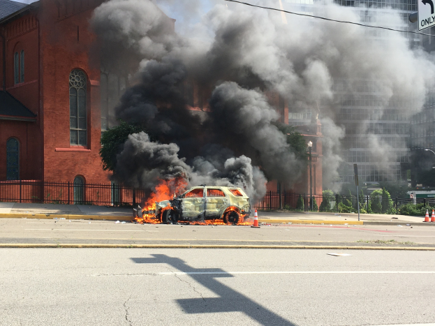 Ein brennendes Auto am Straßenrand mit aufsteigendem Rauch, umgeben von Gebäuden, Bäumen und anderen urbanen Elementen unter einem klaren Himmel.