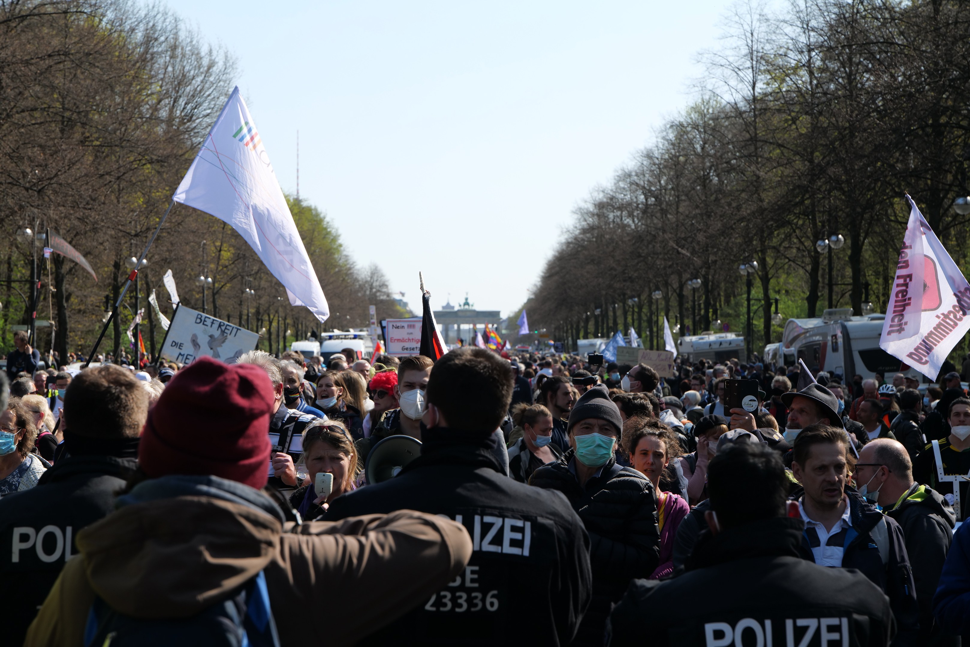 Eine große Gruppe von Menschen vor einer Reihe von Polizeibeamten, einige mit Mützen und Masken, während einer Demonstration in Berlin, Deutschland, mit Schildern, Fahnen, Laternenpfählen, Bäumen, Fahrzeugen und einem Gebäude im Hintergrund.