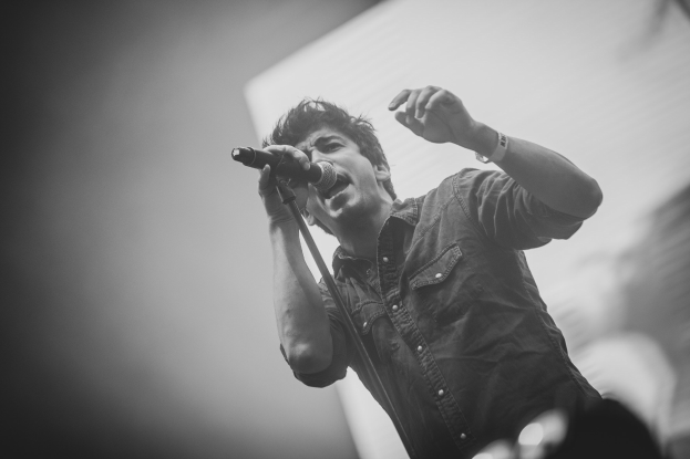 Schwarzes und weißes Foto eines Mannes, der in ein Mikrofon singt, auf der Bühne mit einem unscharfen Gesicht im Hintergrund.