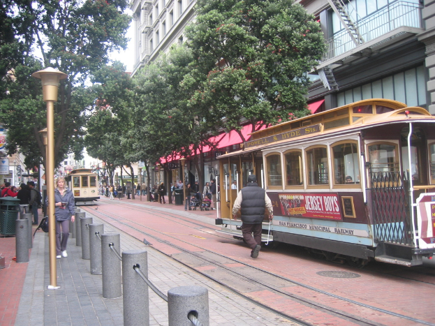 Ein Zug auf Schienen mit Text an der Seite, eine Person in der Nähe, kleine Pfosten mit Seilen links, Straßenlaternen, Bäume und viele Menschen im Hintergrund, ein Müllcontainer links und Gebäude mit Leitern rechts.
