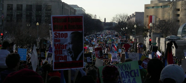 Menschen protestieren auf einer Straße und halten Plakate, mit Gebäuden, Bäumen, Laternenmasten und dem Himmel im Hintergrund.
