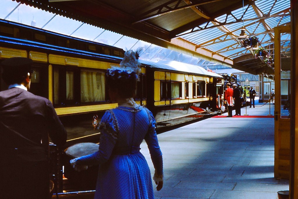 Frau in blauem Kleid neben einem Zug an einem Bahnhof mit Menschen auf dem Bahnsteig.