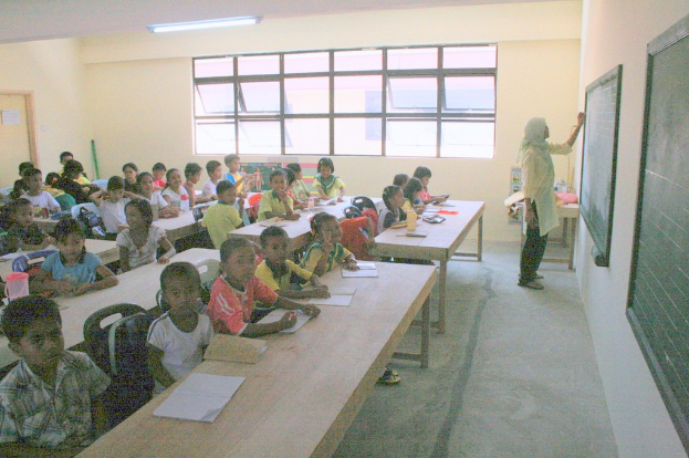 Ein Klassenzimmer mit einer Frau, die rechts an einer Tafel schreibt, Fenstern in der Mitte, einer Lampe an der Decke und Kindern, die an Tischen mit Büchern und Wasserflaschen sitzen.
