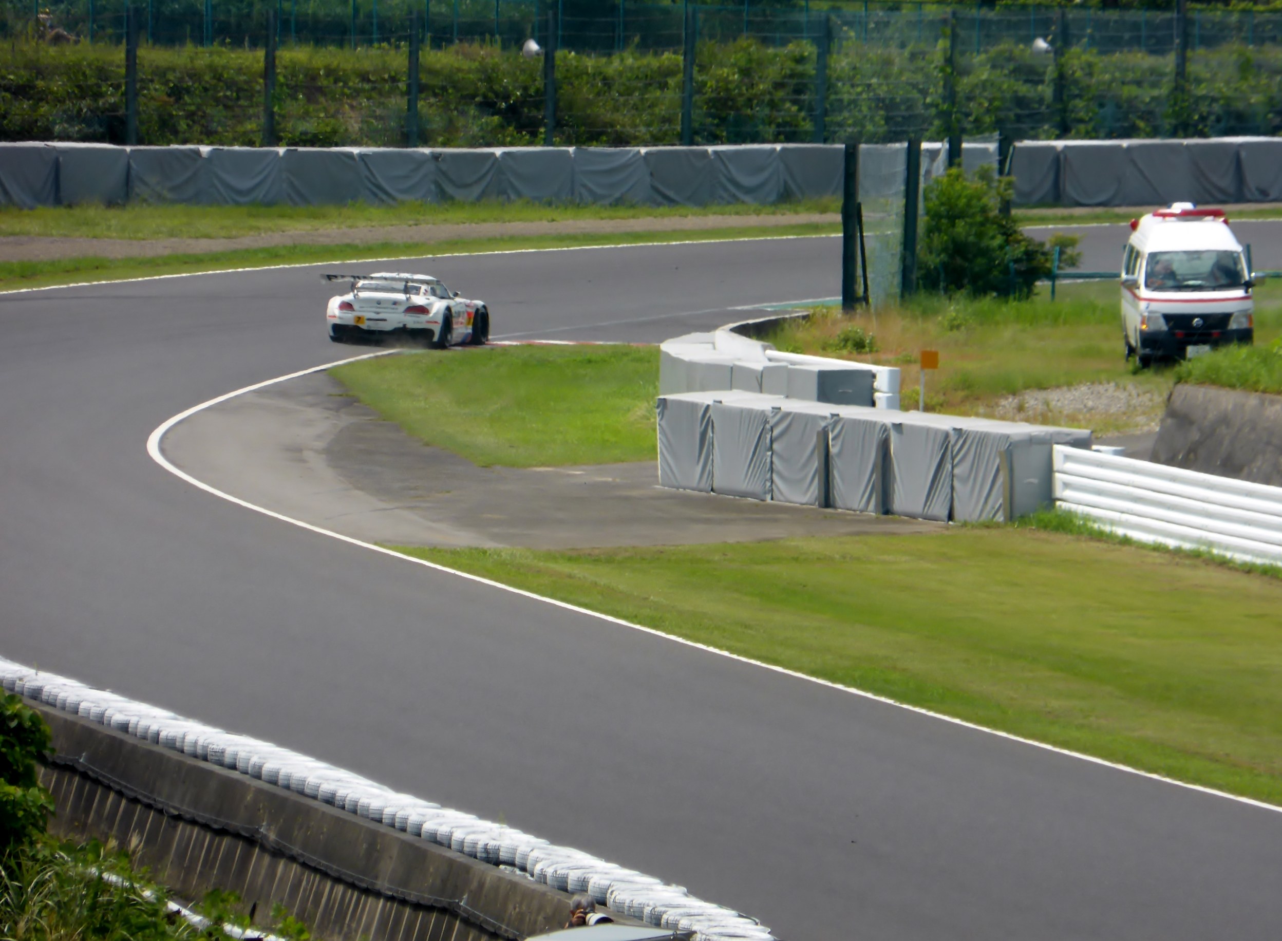 Ein Rennwagen auf einer Rennstrecke mit einem Polizeiwagen im Hintergrund, umgeben von Gras, Pflanzen, Bäumen und Absperrungen, mit einem Fahrzeug auf der rechten Seite und Text unten.