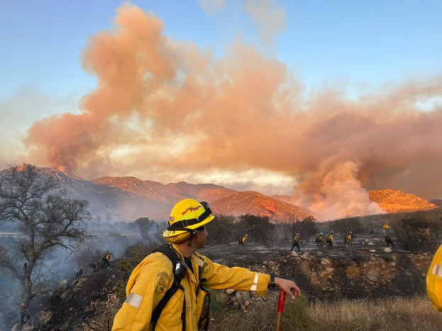 Ein Feuerwehrmann in gelber Jacke und Helm steht vor einer großen Rauchwolke und hält einen Stock, mit Bäumen, Hügeln und einem klaren blauen Himmel im Hintergrund.