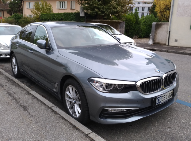 Ein BMW 5er 520d xDrive Luxury Line fährt auf einer Straße mit Gebäuden, Bäumen, Pflanzen und einem klaren blauen Himmel im Hintergrund.