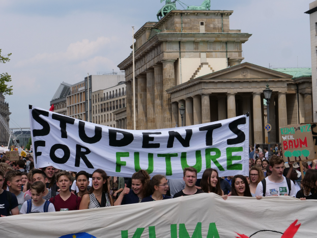 Gruppe von Studenten marschiert in Berlin mit einer bunt bemalten "Students for Future"-Schleife vor einem Hintergrund aus Gebäuden, Bäumen und Himmel.