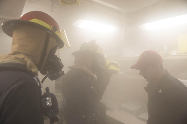 Eine Gruppe von Feuerwehrleuten in Helmen, mit Rauch aus dem Mund, einer hält eine Kamera, in einem Raum mit Lampen und anderen Gegenständen während einer Trainingsrunde.