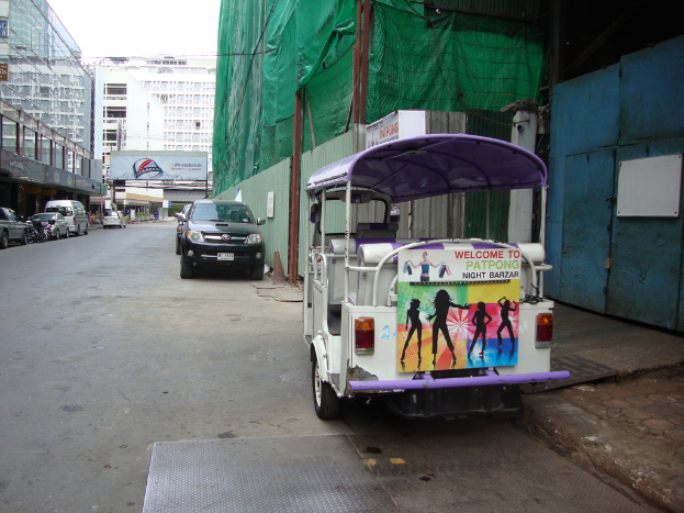 Gebäude mit parkenden Autos, eine Werbetafel und ein bewölkter Himmel, mit einem Auto-Rikscha auf der Seite.