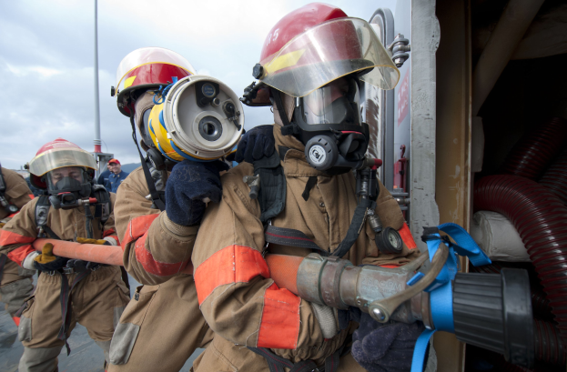 Feuerwehrleute in Schutzausrüstung, einer hält einen Schlauch, mit einer Stange, Himmel, Wolken, Rohren und anderen Gegenständen im Hintergrund.