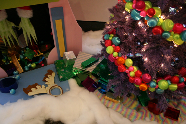 Innenansicht mit einem geschmückten Weihnachtsbaum rechts, Geschenkboxen und Dekorationen in der Mitte, einer Hütte mit Dekorationen oben links und Baumwolle und Wolle unten.