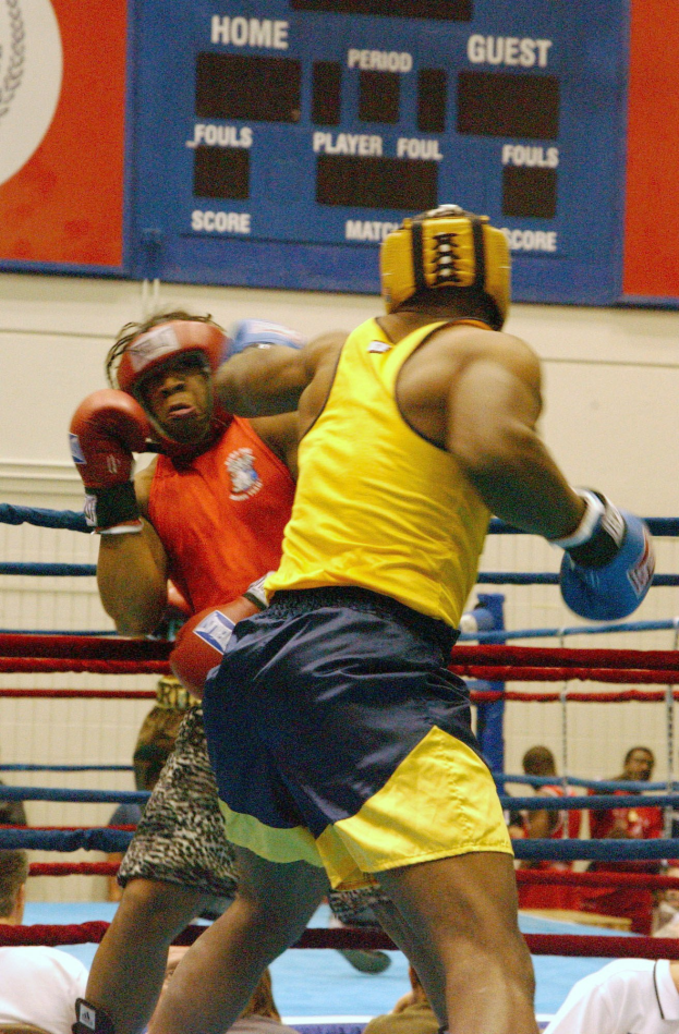 Zwei Boxer, die in einem Boxring kämpfen, umgeben von einer Zuschauermenge, mit einer Wand im Hintergrund, die Text und Logos anzeigt.