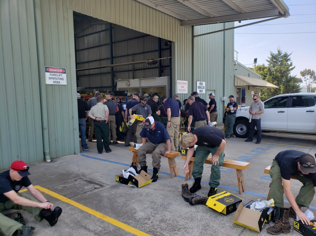 Eine Gruppe von Menschen bei einer Ausbildungssitzung der Nationalgarde, einige sitzen auf Bänken und andere stehen, mit verstreuten Kartons, Schuhen und Gegenständen vor einem Gebäude, vor einer Kulisse aus Fahrzeugen, Bäumen, Gebäuden, Polen, Drähten und einem klaren blauen Himmel.