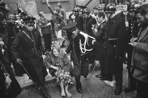 Schwarzes und weißes Bild einer Frau, die von der Polizei eskortiert wird, vor einer Menge mit Helmen und Stöcken, mit einem Gebäude mit Treppe und Geländer im Hintergrund.